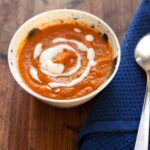 Roasted carrot soup topped with swirls of white cream and a blue napkin and spoon to the side.