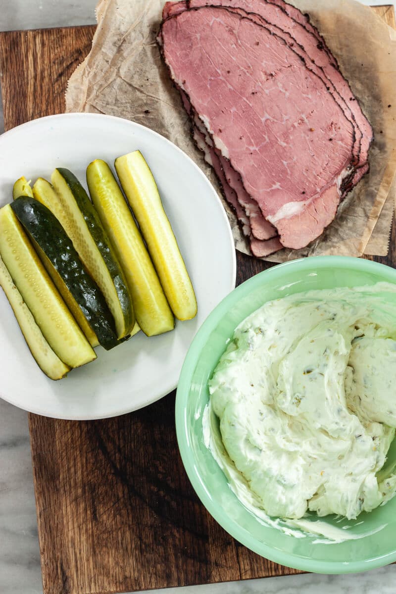 A plate of pickles, some pastrami, and a bowl of seasoned cream cheese on a wooden countertop.