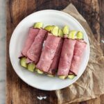 A white plate of pickle rollups on a wooden cutting board with a piece of brown parchment underneath.