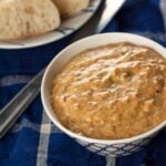 A bowl of roasted pepper dip on a table with a bowl of rolls in the background.