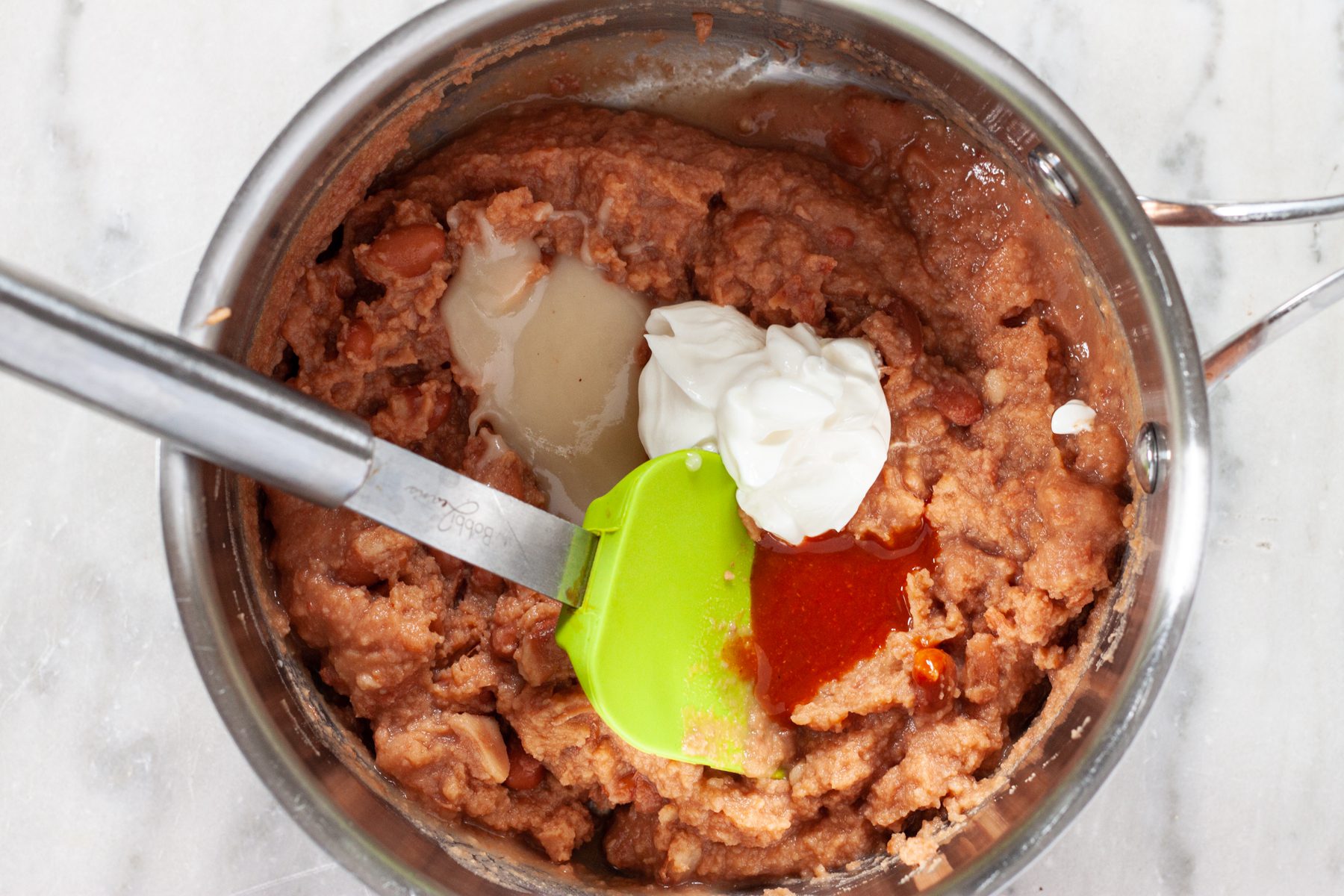 Canned refried beans, sour cream, bacon fat, and hot sauce in a small pot.