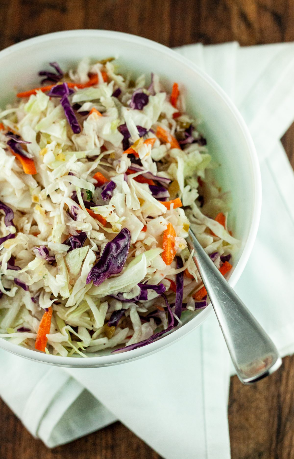 A bowl of oil and vinegar coleslaw dressing tossed with shredded cabbage and carrots and a spoon dipping into it.