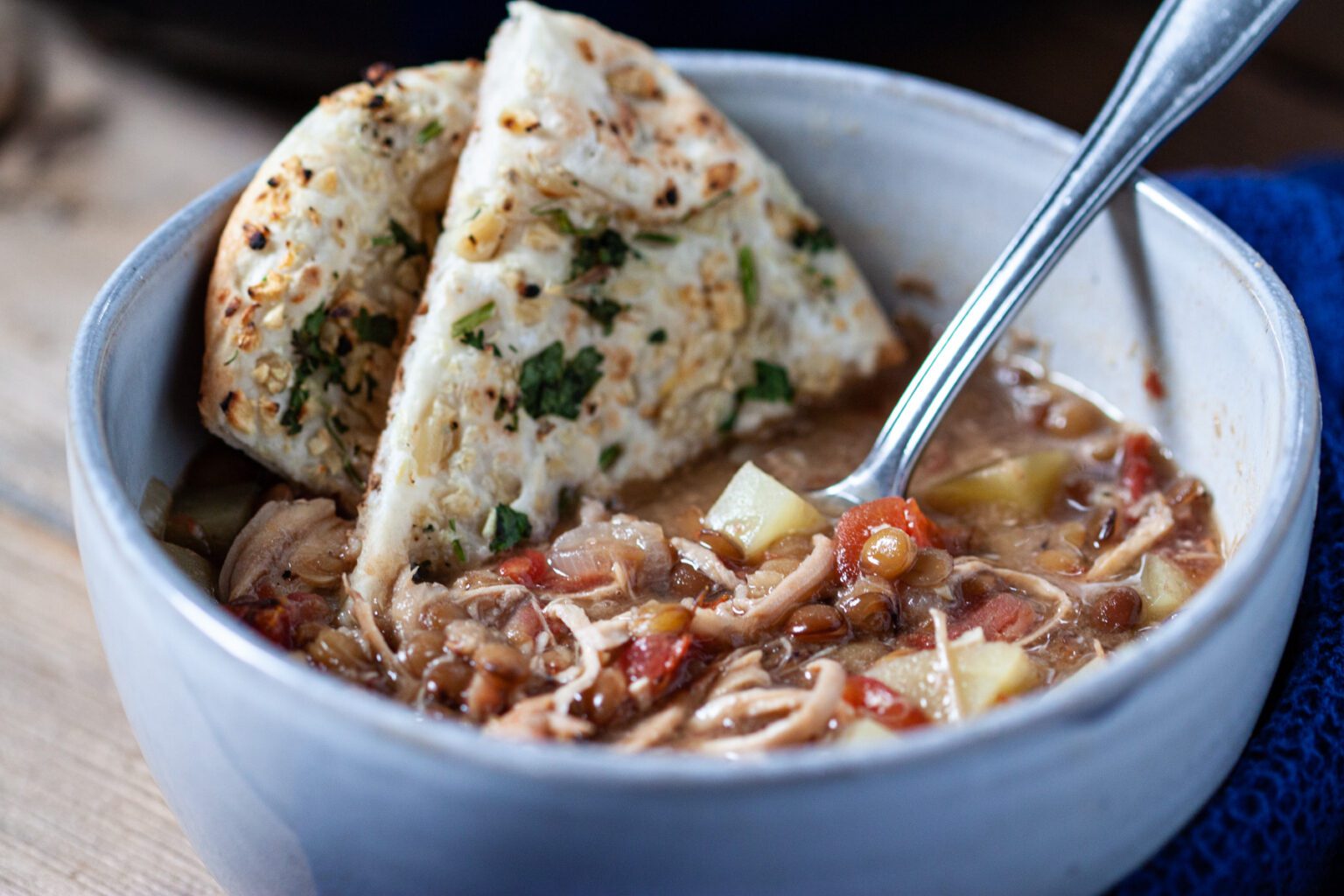 A close-up of a bowl filled with a hearty soup containing vegetables and noodles, garnished with a spoon. Two pieces of herb-infused flatbread are placed on the side of the bowl, partially dipped in the soup, with a blue cloth napkin visible in the background.