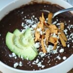 thick black bean soup in a bowl topped with avocado, feta, and tortilla strips in a white bowl.