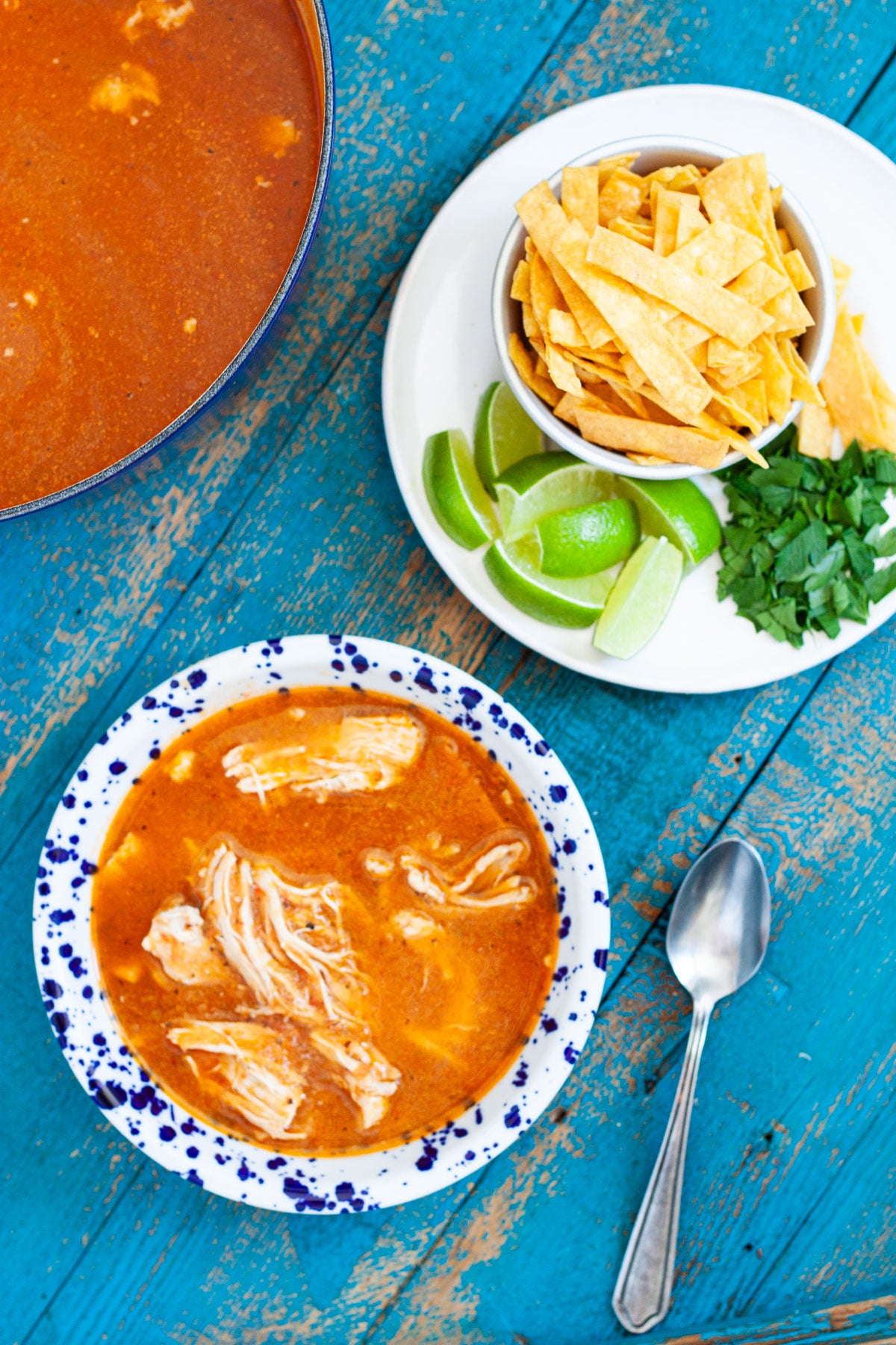 homemade chicken tortilla soup in a bowl sitting next to a plate of garnishes including tortilla strips, lime wedges, and fresh parsley.