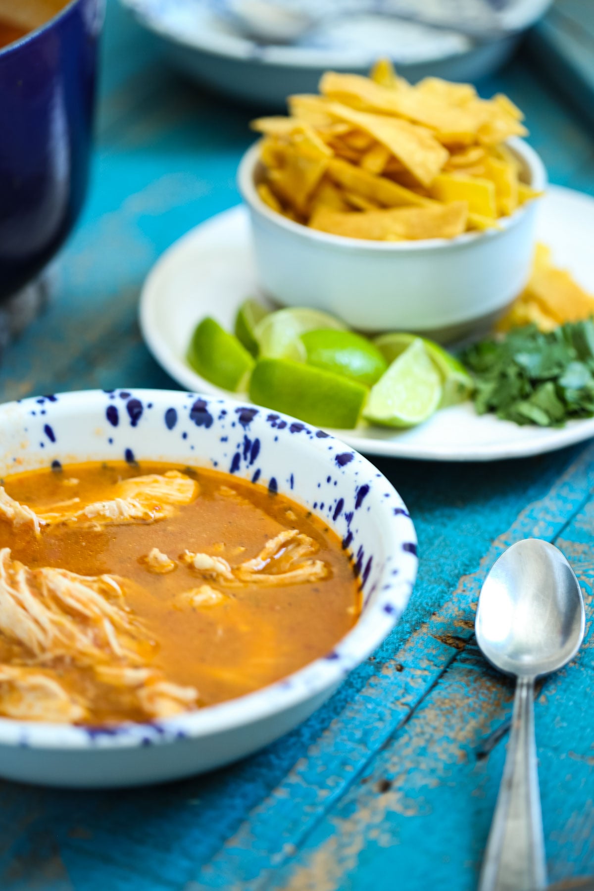 homemade chicken tortilla soup in a bowl sitting next to a plate of garnishes including tortilla strips, lime wedges, and fresh parsley.