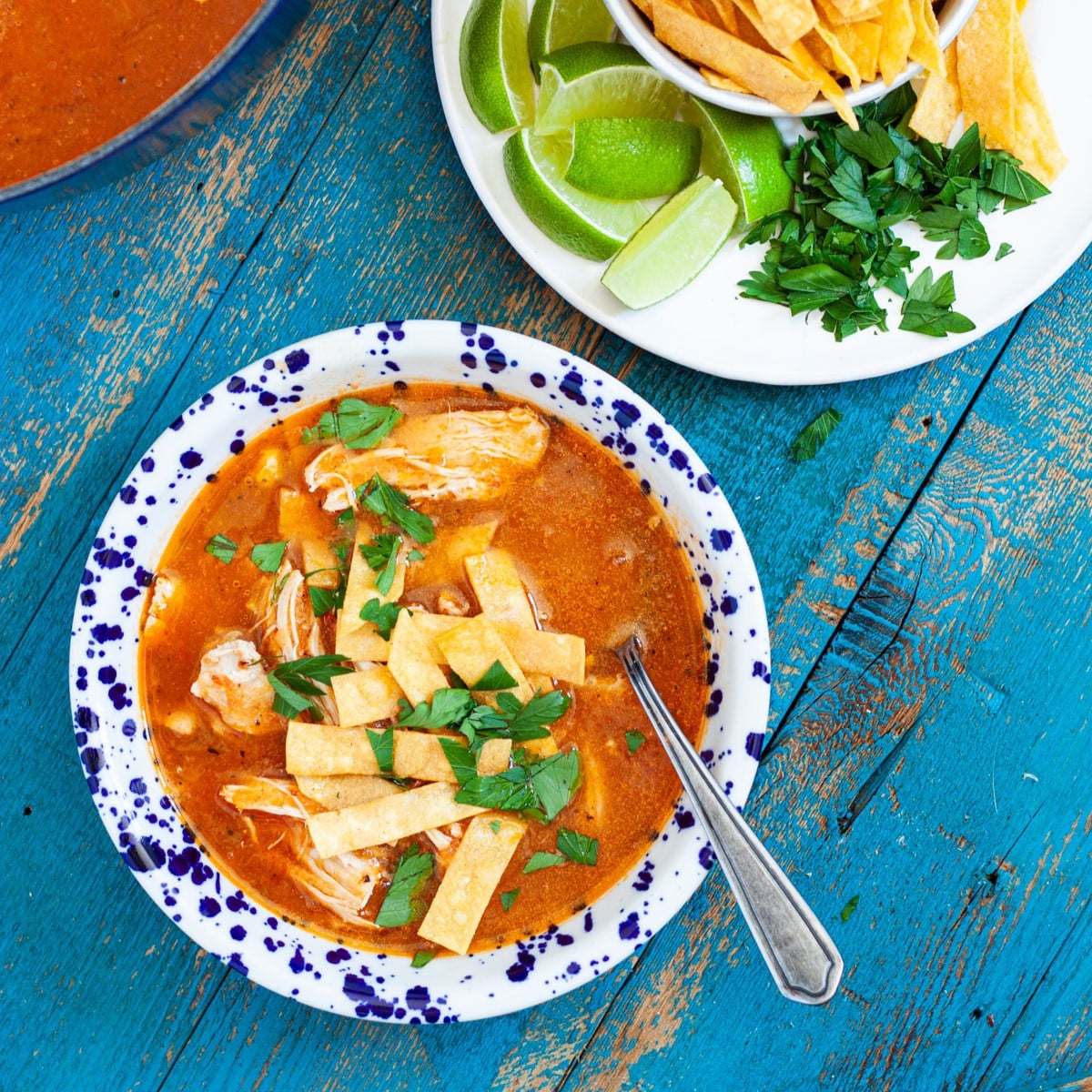 chicken tortilla soup in a bowl with a spoon topped with homemade fried tortilla strips and parsley, sitting next to a plate of garnishes.