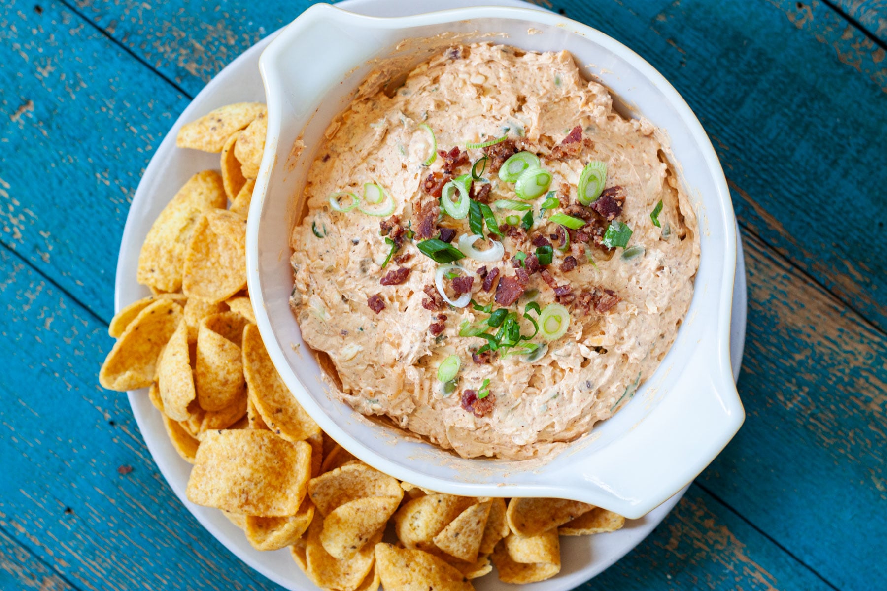 million dollar taco dip garnished with sliced green onion and bacon bits in a bowl next to scoops corn chips on a serving plate.