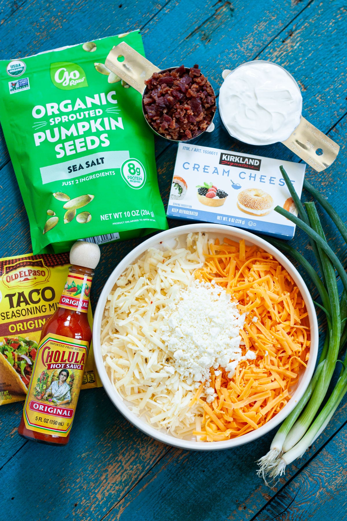 ingredients needed for million dollar taco dip including cream cheese, sour cream, bacon, taco seasoning, hot sauce, and pepitas.