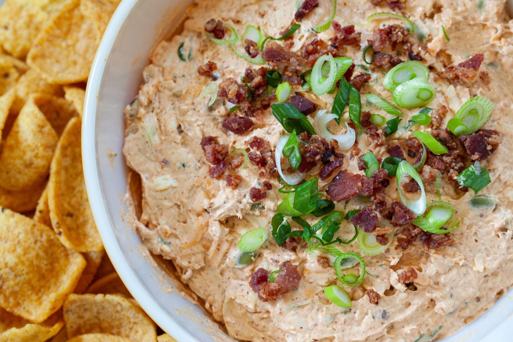 million dollar taco dip garnished with sliced green onion and bacon bits in a bowl next to scoops corn chips.