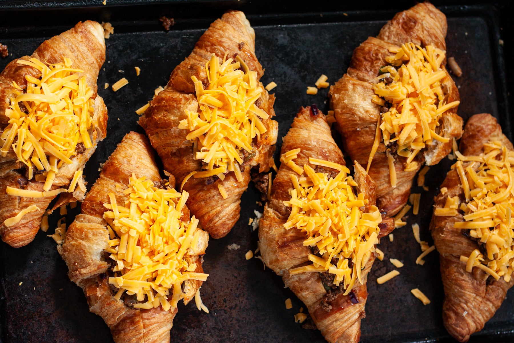 Six assembled croissant tacos topped with shredded cheddar cheese on a baking sheet ready to bake.